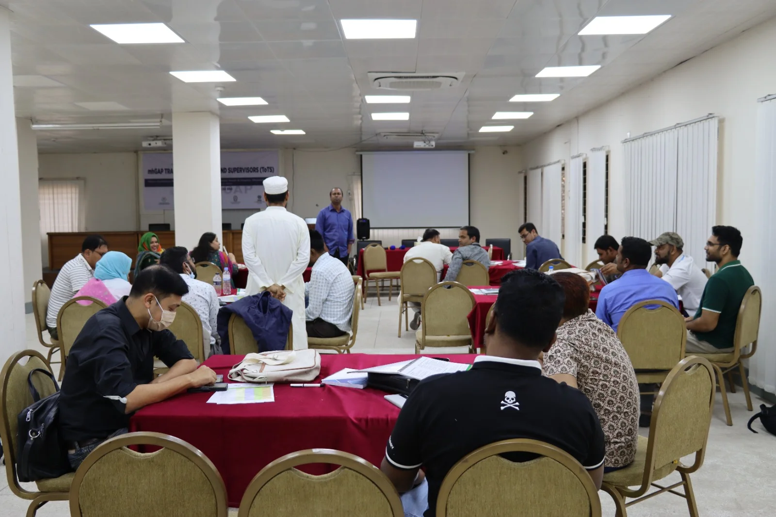 Participants seated at round tables in a training hall, with a banner reading mhGAP Training of Trainers and Supervisors at the front